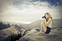 Young woman sitting on a rock on a hill and taking pictures with a camera
