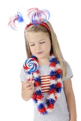 An adorable preschooler dressed to celebrate Independence Day, looking longingly at the red, white and blue swirled lollipop she holds. On a white background.
