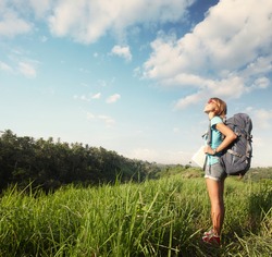 Young smiling woman standing on a green meadow with map and backpack and looking to a blue sky