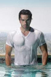 Fashion portrait of a gorgeous male model in soaked wet t-shirt standing in luxurious swimming pool with city background