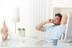 Relaxed man sitting with feet up on desk at home, talking on mobile phone, smiling.