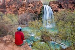 Woman sitting at the edge of a cliff watching Havasu Falls drop into it's turquoise pool. Havasu Canyon, Arizona. Havsupai Reservation.