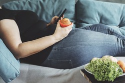 Unhealthy food, mindless snacking, sedentary lifestyle, compulsive overeating. Obese woman laying on sofa with smartphone eating sweets.