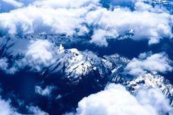 Mountain view from the top through the clouds. The mountain peaks covered with snow. View from airplane. blue tint