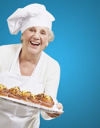 senior woman cook holding a tray with muffins against a blue background