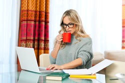 attractive woman with cup of coffee, book and laptop sitting at her home desk and working