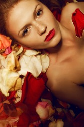 Portrait of a fashionable red-haired (ginger) model with sexy red lips lying on rose petals background. Studio shot