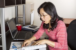 Mature Asian woman surprised while holding income tax tables booklet with computer, coffee cup and calculator on desk