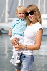 Young white blonde woman holding her little blond son in hands outdoor.Happy family walking on waterfront with yachts on background.Young adult woman holding child.Happy mother with cute kid in summer