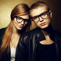 Portrait of gorgeous red-haired fashion twins in black clothes wearing trendy glasses and posing over golden background together. Perfect hair. Natural make-up. Perfect skin. Studio shot.