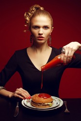 Unhealthy eating. Junk food concept. Portrait of fashionable young woman holding bottle with tomato sauce, pouring it and going to eat burger. Perfect hair, skin, make-up and manicure. Studio shot