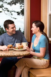 An attractive couple talking over breakfast by a large window, with a picturesque background, at home. Vertically framed shot.