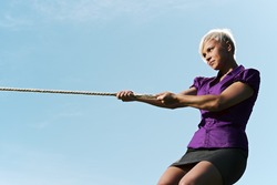 resolute business woman pulling rope against blue sky, symbol of power and determination. Copy space, side view