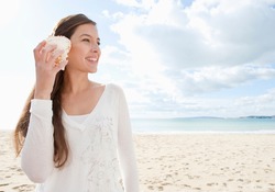 Young attractive healthy woman on a white sand beach holding a sea shell against her ear and listening to the sound of waves smiling, during a vacation trip away.