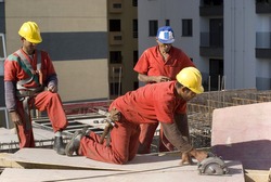 One worker is making a cut a on a sheet board with a circular saw. Their are two other workers standing behind him. Horizontally framed shot.