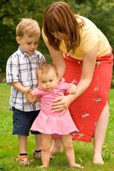 Young mother and her son helping baby daughter to walk in the garden.