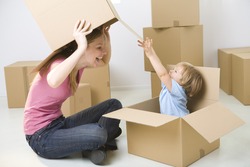 A woman with young girl sitting between cardboard boxes. Happy woman holding box over head and looking at young girl. Young girl sitting in box.