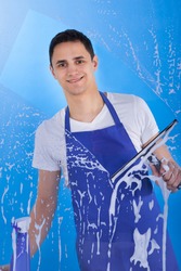 Portrait of young male servant cleaning glass with squeegee over blue background