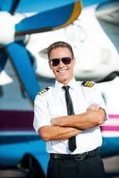 My job is my passion. Confident male pilot in uniform keeping arms crossed and smiling while standing in front of the airplane