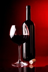 Red wine glass and wine bottle and cork bottle stopper. Placed on glass table with small mirror reflection. Red background light.