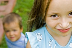 A little girl looking straight at the camera and a little boy sitting on the ground in the background.