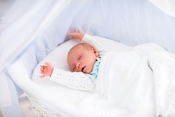 Newborn baby boy waking up on a sunny morning in white bed. New born child sleeping and stretching in moses basket under knitted blanket. Children sleep. Bedding for kids. Infant napping in bed.