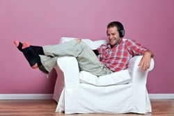 Male in casual clothing relaxing on an armchair wearing headphones whilst watching and listening to a music video on his media player.