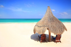 Two beach chairs under the shade of a grass umbrella overlooking a stunning tourist resort beach