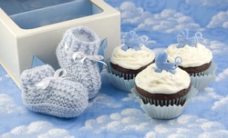 A pair of crocheted blue baby booties, a gift box and delicious chocolate cupcakes with vanilla frosted and baby carriage decoration, baby shower gifts, vertical with copy space