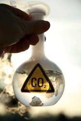 a Scientist holds a 500ml beaker filled with CO2 gainst the blue sky, with a truck in the background representing carbon emmissions from vechiles