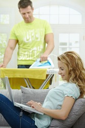 Wife sitting on couch using laptop, husband ironing in the background, smiling. Selective focus on woman.