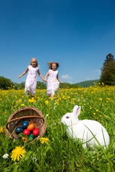 Easter bunny on a beautiful spring meadow with dandelions in front of a basket with Easter eggs; children in the background coming on an Egg hunt