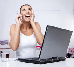young excited woman working at home on her laptop computer while talking on a mobile phone