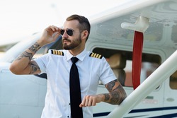 Cool young adult pilot with tattoos in sunglasses posing at private motor airplane on runway near hangar.