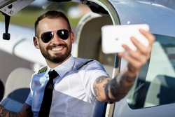 Cool young pilot take a selfie with his smart phone sitting in air craft cabin. Handsome man with tattoos