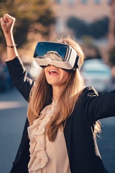 Excited young beautiful girl gesture testing virtual reality 3D video glasses VR headset dressed in a office outfit impressed by augmented reality on the street and beautiful autumn sun light colors