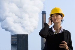 A beautiful dark haired woman wearing a hard hat and talking on her mobile phone while standing in front of a factory pumping out pollution from its chimneys