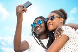 Close up portrait of two diverse african teen girls wearing sun glasses taking self portrait with phone against blue sky.