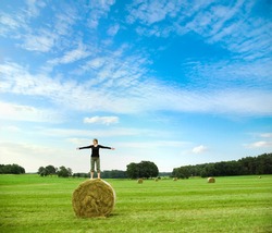girl standing on hay bale spreading her arms in front of a beautiful landscape with bright blue sky