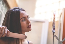 Stunning young woman straightening her hair with an iron - Beautiful woman with makeup getting ready in the bathroom
