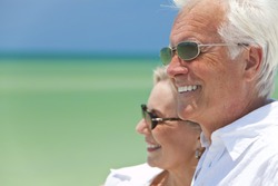 Happy senior man and woman couple together looking out to sea on a deserted tropical beach with bright clear blue sky