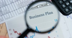 A hand with a magnifying glass passes over a financial newspaper and financial bag with a business plan. The lens magnifies the word "Business Plan". Concept: economy, finance, crisis, business plan,