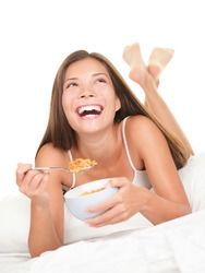 Woman eating breakfast in bed. Happy woman enjoying cornflakes cereals in the bed in the morning. Beautiful mixed race Chinese Asian / Caucasian female model lying in bed isolated on white background