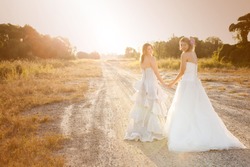 Attractive young bride and bridesmaid in formal attire looking over their shoulders at the camera. They are walking on a country road. Horizontal shot.