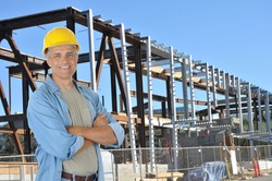 Construction worker with arms crossed on job site standing in front of new building frame. Horizontal format with man smiling at camera.