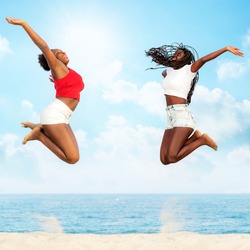 Full length action portrait of two african girlfriends jumping together on beach. Girls screaming with hands in air.