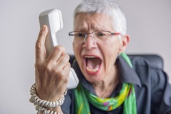 Angry, enraged senior woman yelling at a landline office phone, unhappy with customer service provided by the agent on the other side