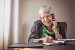 Happy senior old business woman writing down orders, talking on her cell phone