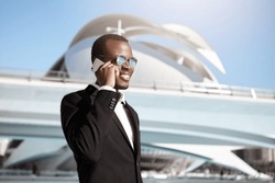 Successful dark-skinned businessman wearing elegant black suit and round sunglasses having pleased smile while speaking over smart phone with his business partner isolated over big city background