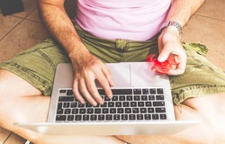 Young adult man seated on a floor, working on laptop and playing with a red hand spinner fidget.